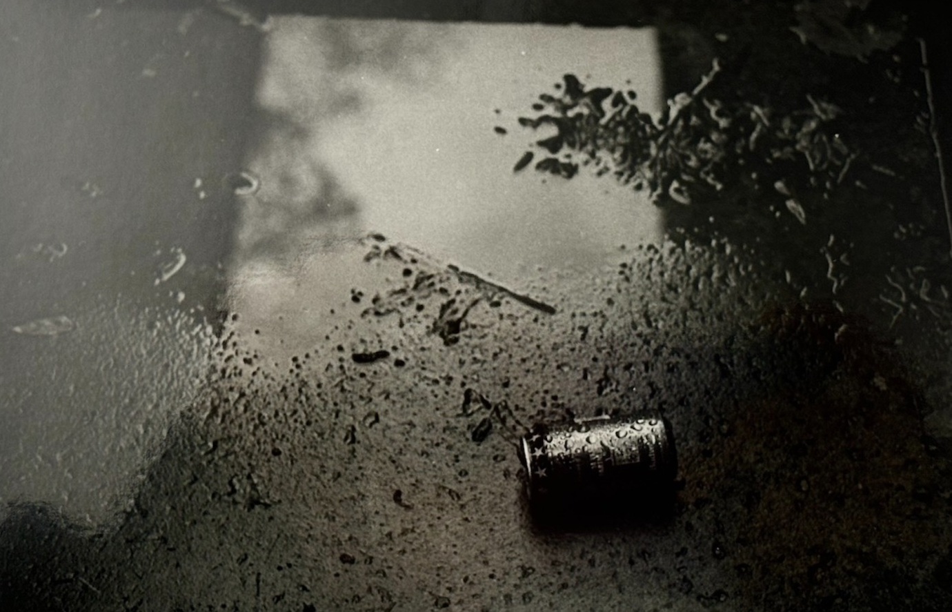 A crushed can on rain-darkened pavement beside a reflection